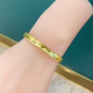 May include: A gold bangle bracelet with a textured, geometric design. The bracelet is worn on a wrist and has a bright, shiny appearance. The background is a solid teal colour.