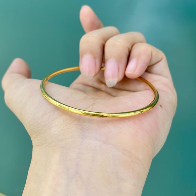 K&ouml;nnte beinhalten: Ein goldfarbenes Armreif-Armband. Das Armband ist ein einfaches, d&uuml;nnes Band mit glatter, polierter Oberfl&auml;che. Der Armreif wird in einer Hand vor einem t&uuml;rkisfarbenen Hintergrund gehalten.