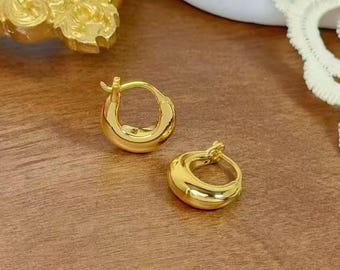 Puede incluir: Un par de pendientes de aro pequeños, de color dorado. Los pendientes tienen forma de media luna redondeada y un acabado pulido. Los pendientes se muestran sobre una superficie marrón, con un elemento decorativo dorado en el fondo.