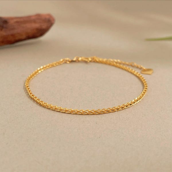 May include: A delicate gold bracelet with a woven chain design. The bracelet is a bright gold color and features a small heart-shaped charm at the clasp. The bracelet is shown on a neutral background.