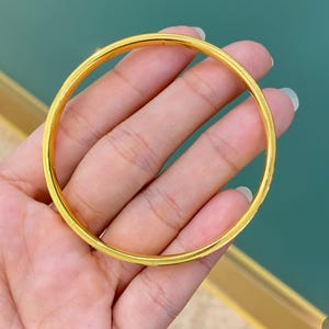 May include: A gold-coloured bangle bracelet with a textured, ridged design. The bracelet is circular and appears to be made of metal. The bangle is held in a hand against a teal background.
