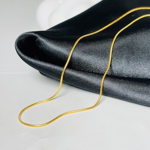 Puede incluir: Un collar de cadena de serpiente fino de color dorado con una textura suave y flexible. La cadena presenta un diseño de eslabones continuos y uniformes que crea una apariencia metálica elegante, reflejando la luz en su superficie redondeada.