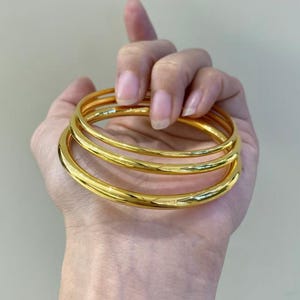 K&ouml;nnte beinhalten: Eine Hand h&auml;lt drei goldfarbene Armreifen. Die Armreifen sind glatt und rund und reflektieren das Licht, wodurch sie gl&auml;nzend wirken. Das Schmuckst&uuml;ck ist ein schlichtes, elegantes Accessoire.