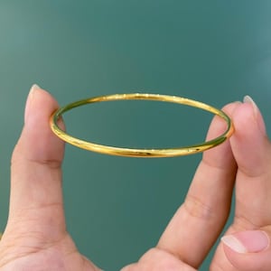 K&ouml;nnte beinhalten: Ein goldener Armreif, der zwischen zwei Fingern gehalten wird. Der Armreif ist ein durchgehender, ununterbrochener Kreis mit einer glatten, reflektierenden Oberfl&auml;che. Der Hintergrund ist einfarbig t&uuml;rkis.