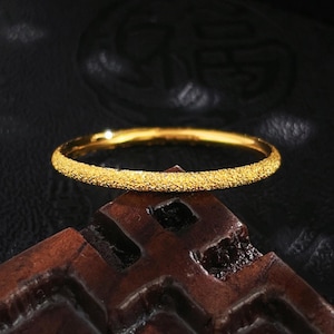 May include: A thin, circular gold ring featuring a textured, sandblasted finish that creates a sparkling, granular surface appearance. The band is uniform in width and maintains a consistent, bright yellow-gold color throughout its entire circumference.