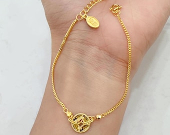 Puede incluir: Pulsera dorada con una cadena delicada y un dije circular con detalles intrincados. El dije tiene un diseño de filigrana con acentos negros y amarillos. La pulsera se muestra en una muñeca, mostrando su longitud ajustable.