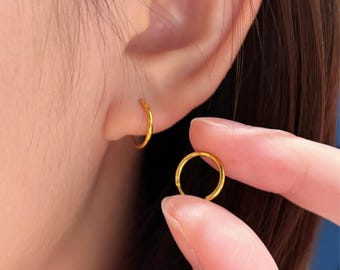 Puede incluir: Primer plano de una oreja con un pequeño pendiente de aro dorado. Otro pendiente de aro dorado se sostiene con los dedos. Los pendientes son simples, circulares y tienen un acabado brillante. El tono de piel es claro.