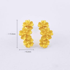 Puede incluir: Un par de pendientes de color dorado, cada uno con tres formas de flores texturizadas dispuestas en un grupo vertical. Los pendientes miden aproximadamente 15,4 mm de alto y 8,2 mm de ancho, seg&uacute;n indican los marcadores de medida.