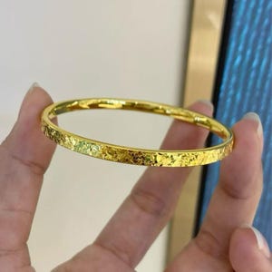 May include: A gold-colored bangle bracelet with a textured surface. The bracelet has a hammered design, reflecting light. The jewelry is a simple, elegant accessory.