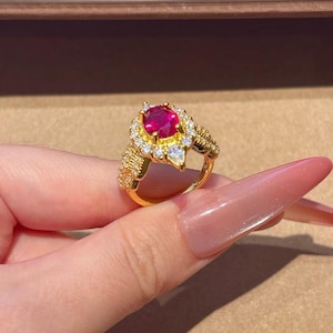 Könnte beinhalten: Ein goldener Ring mit einem zentralen, oval geschliffenen, leuchtend roten Edelstein, der von einem Kranz aus kleinen, klaren Steinen umgeben ist. Die Ringschiene ist pavéartig texturiert und an den Seiten mit weiteren klaren Steinen besetzt.