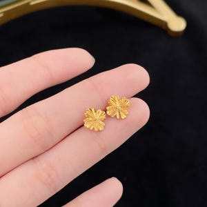 May include: A pair of small, gold-colored clover-shaped stud earrings. The earrings have a detailed, textured surface and are displayed against a dark background.