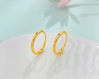 May include: A pair of small, gold-colored hoop earrings. The earrings have a textured surface and are displayed on a white, wavy surface against a soft blue and pink background. The earrings are a simple, elegant design.