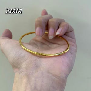 K&ouml;nnte beinhalten: Ein goldfarbenes Armreif-Armband, 2 mm dick, wird in einer Hand gehalten. Das Armband hat eine glatte, polierte Oberfl&auml;che und ein schlichtes, elegantes Design. Der Text "2MM" befindet sich in der oberen linken Ecke.