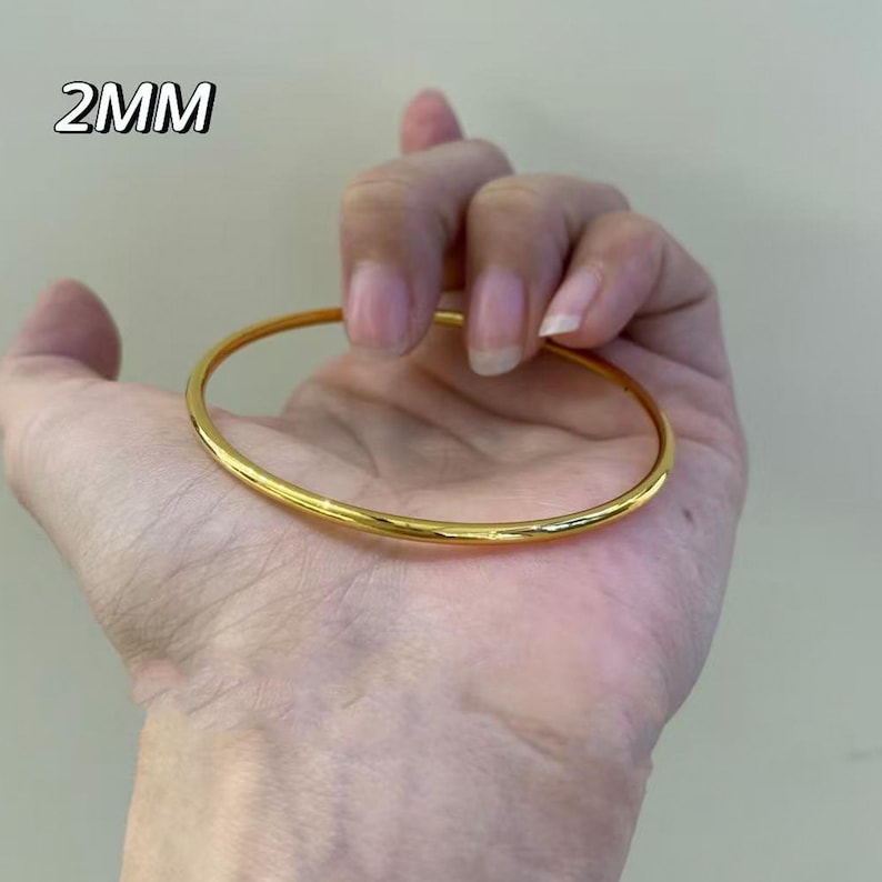 K&ouml;nnte beinhalten: Ein goldfarbenes Armreif-Armband, 2 mm dick, wird in einer Hand gehalten. Das Armband hat eine glatte, polierte Oberfl&auml;che und ein schlichtes, elegantes Design. Der Text "2MM" befindet sich in der oberen linken Ecke.