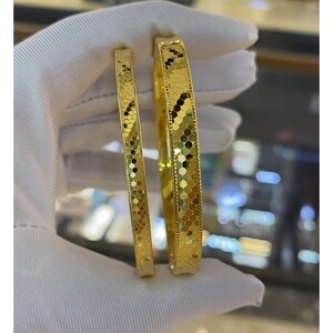 May include: Two gold bangles with textured surfaces are shown. The bangles feature a repeating pattern of small, connected shapes. The larger bangle is wider than the smaller one. The jewellery is held against a white background.