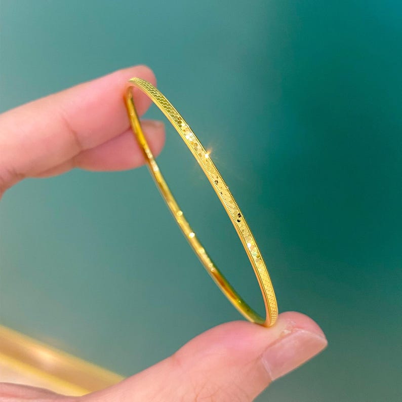 May include: A slender, gold-coloured bangle bracelet with a textured surface. The bracelet is held against a teal background, highlighting its delicate design and subtle sparkle. The bangle is a simple, elegant piece of jewellery.