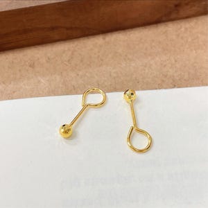 Puede incluir: Un par de pendientes de oro, cada uno con un poste fino y recto conectado a un pequeño bucle circular en un extremo y una cuenta esférica texturizada en el otro. El metal dorado tiene un acabado reflectante y las cuentas esféricas tienen una superficie facetada que capta la luz, creando una apariencia brillante y distintiva para estas piezas de joyería.