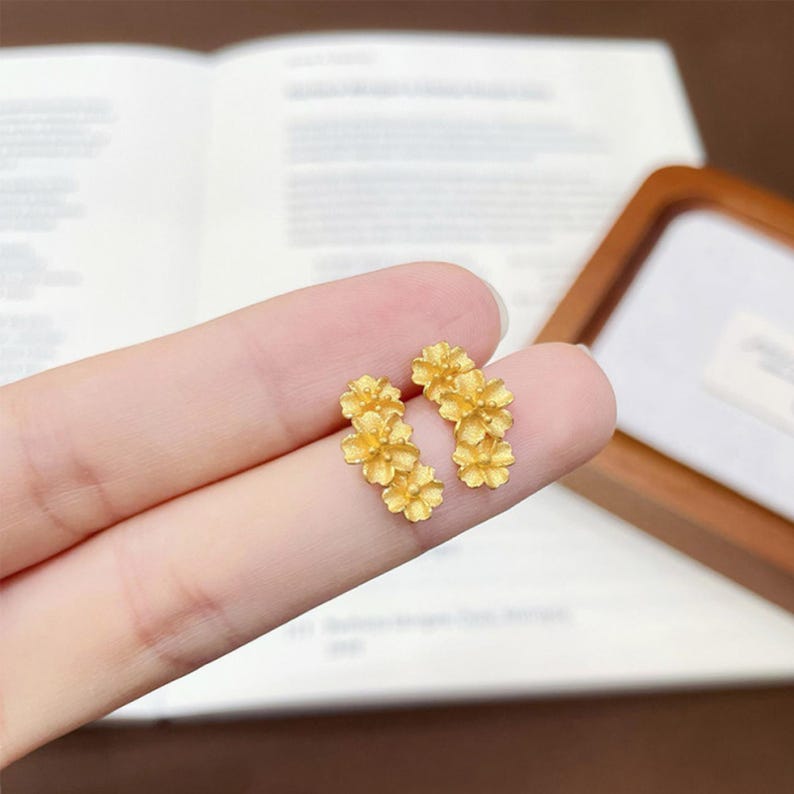 Puede incluir: Un par de pendientes de bot&oacute;n en tono dorado, cada uno con un grupo de tres peque&ntilde;as flores de cinco p&eacute;talos con un acabado mate texturizado. El dise&ntilde;o floral est&aacute; dispuesto en una l&iacute;nea vertical, creando un aspecto bot&aacute;nico detallado.
