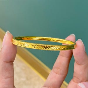 May include: A gold bangle bracelet with a textured surface and a repeating pattern of small shapes. The bracelet is held between two fingers against a teal and green background. The bangle is approximately 6.35 cm in diameter.