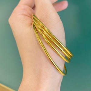 K&ouml;nnte beinhalten: Eine Hand h&auml;lt mehrere d&uuml;nne, goldfarbene Armreifen. Die Armreifen sind glatt und gl&auml;nzend und reflektieren das Licht. Der Hintergrund ist einfarbig t&uuml;rkis und bildet einen Kontrast.