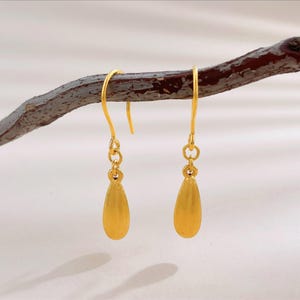 Op de afbeelding: Een paar goudkleurige oorbellen met een gladde, druppelvormige hanger die aan een gebogen haakje hangt. Elke oorbel is verbonden door een kleine ronde ring en heeft een matte afwerking op het metalen oppervlak van de druppelvorm.