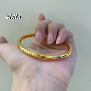 K&ouml;nnte beinhalten: Ein gl&auml;nzendes, goldfarbenes Armreif-Armband. Das Armband ist 4 mm dick und hat ein glattes, abgerundetes Design. Der Text "4MM" ist sichtbar. Das Armband wird in einer Hand gehalten.