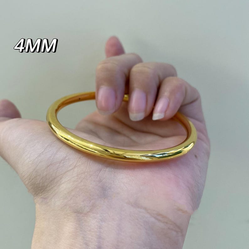 K&ouml;nnte beinhalten: Ein gl&auml;nzendes, goldfarbenes Armreif-Armband. Das Armband ist 4 mm dick und hat ein glattes, abgerundetes Design. Der Text "4MM" ist sichtbar. Das Armband wird in einer Hand gehalten.