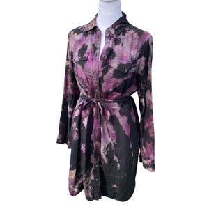 May include: A long-sleeved shirt dress with a tie-waist, featuring a tie-dye pattern in shades of black, purple, and white. The dress has a collared neckline, button-down front, and two chest pockets. The dress is displayed on a mannequin.