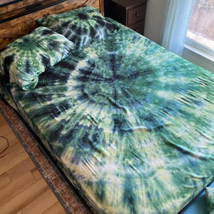 May include: A bed with a tie-dye pattern in shades of green, blue, and black. The bedding includes a fitted sheet and two pillowcases. The bed frame is dark brown with a metal headboard.