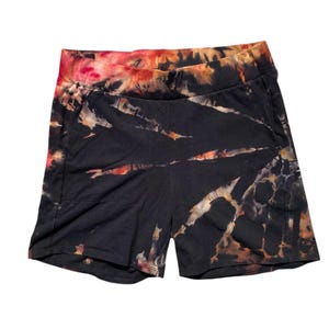 May include: Black tie-dye shorts with a wide waistband. The shorts feature a unique pattern of orange, red, and cream-coloured streaks against a black background. The shorts have pockets and are made of a soft, comfortable material.