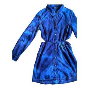 May include: A long-sleeved, button-down denim dress with a tie-dye pattern in shades of blue. The dress features a collared neckline, pockets, and cut-out details at the waist. White stitching accents the seams.