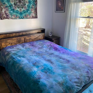 May include: A bed with a tie-dye duvet cover in shades of blue, purple, and brown. A matching tapestry hangs above the bed, and a framed picture is on the wall. The bed frame is dark wood.