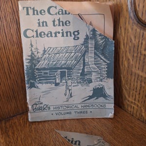 May include: A vintage book titled "The Cabin in the Clearing" with a black and white illustration of a cabin and figures. The book is part of "Jim Baker's Historical Handbooks, Volume Three."