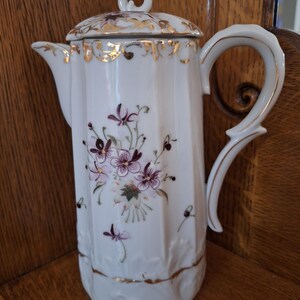 May include: White porcelain coffee pot with a lid, adorned with purple floral designs and gold trim. The pot features a curved handle and a spout for pouring. A decorative piece for serving coffee or tea.