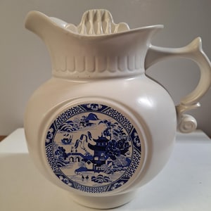 May include: White ceramic pitcher with a blue and white Willow pattern design. The pitcher has a spout, handle, and a built-in juicer. The round body features a circular blue and white landscape scene. A decorative kitchenware item.