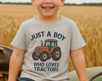 May include: A young child wearing a gray t-shirt with a tractor graphic and the text "JUST A BOY WHO LOVES TRACTORS." The child is smiling in front of a field of wheat.