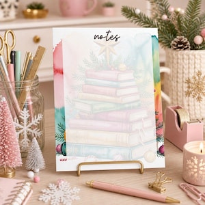 May include: A rectangular notepad featuring the word notes at the top, decorated with a watercolor illustration of a stack of books topped with a star, pine branches, and ornaments. The notepad is displayed on a gold metal stand.