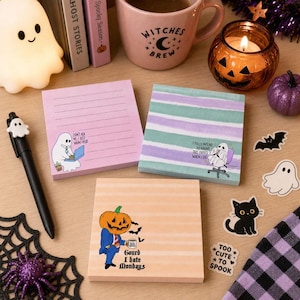 May include: Three square sticky note pads featuring Halloween themes. One pink pad with lines and a ghost at a laptop, one teal and purple striped pad with a ghost in an office chair, and one orange striped pad with a pumpkin-headed figure in a suit.