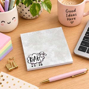 May include: A square notepad with a light gray textured surface featuring a cartoon sheep illustration in the bottom left corner. The sheep has black outlines and the word BAH written in black capital letters inside its fluffy white body.