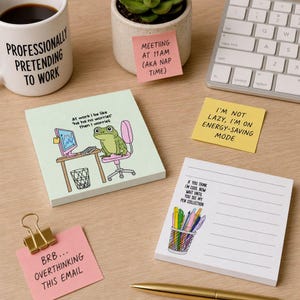 May include: A collection of stationery items including a white mug with black text, a light green notepad with a frog illustration, a yellow sticky note with black text, a pink sticky note with black text, and a white lined notepad with a pen cup graphic.