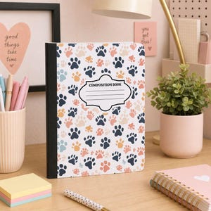 May include: A composition book featuring a repeating pattern of paw prints in shades of dark blue, light blue, peach, and tan. The cover has a central white label with a black border and the text COMPOSITION BOOK with lines for writing.