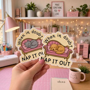 May include: Two die-cut stickers featuring a pink sofa with a sleeping animal. One sticker shows a gray cat, the other a golden dog. Both stickers have the text "When in doubt NAP IT OUT" in black lettering against a light yellow background.