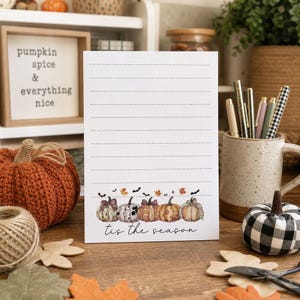 May include: A white rectangular stationery sheet with horizontal lines, featuring a bottom border of watercolor-style pumpkins, autumn leaves, and small black bats. The text 'tis the season' is printed in black script below the pumpkins.
