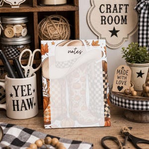 May include: A rectangular notepad featuring a central white area for writing labeled notes, decorated with a graphic of a cowboy hat and boots patterned with pumpkins and checkered designs, surrounded by autumn leaves and small stars on a white background.