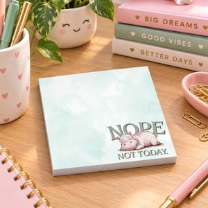 May include: A square notepad with a light blue watercolor background featuring a cartoon illustration of a pink hippopotamus lying down. The text NOPE NOT TODAY is printed in a dark gray serif font above the hippo on the bottom right corner.