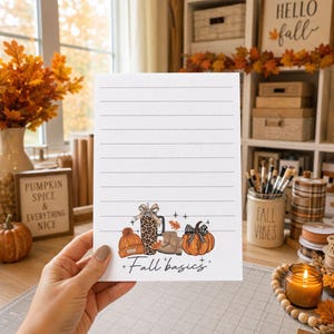 May include: A white lined notepad featuring a colorful illustration at the bottom with an orange knit beanie, a leopard print tumbler with a bow, tan boots, and an orange pumpkin with a plaid bow, accompanied by the text Fall basics.