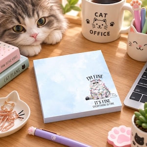 May include: A square notepad with a light blue watercolor background featuring a fluffy cat wrapped in colorful string lights. The text reads I'M FINE IT'S FINE EVERYTHING IS FINE. The notepad is positioned on a wooden desk surface.