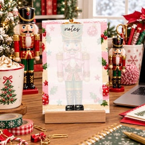 May include: A rectangular notepad featuring a nutcracker illustration and the word notes at the top, held upright by a wooden stand. The notepad has a pink and cream background decorated with red ornaments, green holly leaves, and small stars.