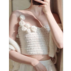 May include: Cream-colored crochet crop top with delicate rose embellishments. The top features thin straps and a fitted design. A matching cream-colored cardigan is draped over the shoulders, creating a soft, textured look. The top is paired with a white skirt.
