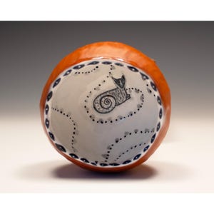 May include: A ceramic bowl with a white interior featuring a black cat illustration. The bowl has a light orange exterior and a decorative border of blue and black dots. The cat is curled up with a swirled tail.